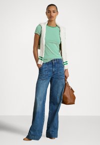 Žena v zeleno-bielom pruhovanom tričku, s krémovým svetrom prehodeným cez plecia, v širokých modrých džínsach, drží hnedú koženú kabelku.