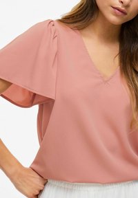 Blusa rosa pallido con maniche corte a sbuffo, scollatura a V e texture di tessuto morbido. Presenta una leggera arricciatura ai punti spalla.