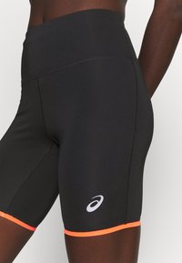 Black athletic shorts made of stretchy material featuring an orange accent at the hem and a white logo on the side. High-waisted design.
