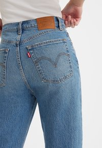 Jean en denim bleu clair avec une taille haute, poches arrière avec des détails de couture et une étiquette de marque en cuir brun.