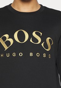 Maglia nera a maniche lunghe con grande scritta dorata "BOSS" e scritta più piccola "HUGO BOSS", ricamate con una finitura testurizzata.