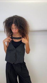 Schwarze Weste mit Nadelstreifen und Knöpfen, über einem schwarzen Tanktop getragen. Das Model hat lockiges Haar und passt die Weste an, um ihr Design zu zeigen.
