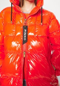 Czerwona kurtka puchowa o błyszczącym wykończeniu, z zamkiem z przodu, regulowanym kapturem i czarną metką marki "PINKO". Wyróżniający się pikowanym wzorem.