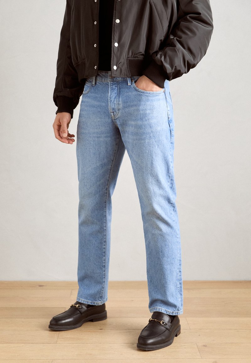 Jeans en denim bleu clair avec une coupe droite, portés avec une veste bomber en nylon noire et des mocassins foncés avec des détails en métal doré.