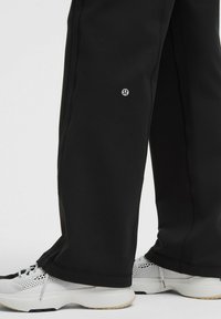 Schwarze, weit geschnittene Hosen mit einem kleinen Logo über dem Knöchel, kombiniert mit weißen, atmungsaktiven Sportschuhen auf einem weißen Hintergrund.