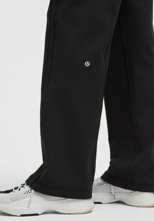 Pantaloni neri a gamba larga con un piccolo logo sopra la caviglia abbinati a sneakers da atletica in mesh bianco su uno sfondo bianco.