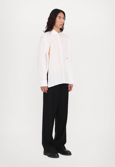 Chemise blanche à manches longues avec un détail découpé, assortie à un pantalon noir à jambes larges et des chaussures noires à enfiler, présentant un tissu texturé et un logo discret.