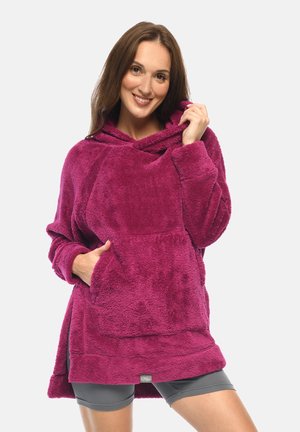 Frau lächelt, trägt einen kuscheligen, lila, übergroßen Hoodie mit Fronttasche und graue Shorts, hält mit einer Hand die Kapuze.