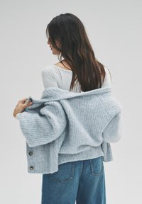 Gaiši zils adīts kardigans ar reljefu rakstu un lielām pogām. Valkāts virs pelēkas garroku blūzes un kombinēts ar džinsa biksēm.