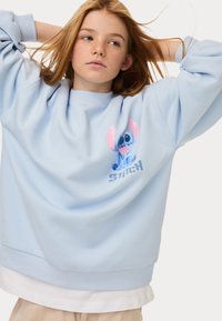 Felpa azzurra chiara in misto cotone, con una grafica frontale di Stitch e la parola "STITCH" in un blu più scuro. Tessuto morbido, vestibilità rilassata.