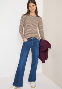Langärmliges Oberteil in Braun- und Cremestreifen, kombiniert mit weiten blauen Jeans. Das Model hält eine kastanienbraune Jacke und trägt weiße Sneaker.