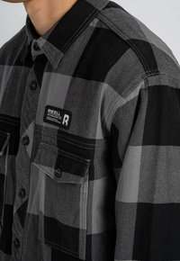 Schwarz-graues karierter Flanellhemd mit Knopfleiste, zwei Brusttaschen und einem Etikett auf der linken Tasche. Strukturierter Stoff.