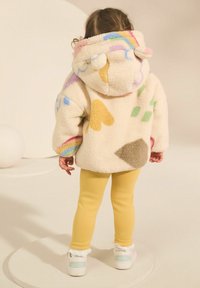 Veste en polaire de couleur crème avec des motifs abstraits colorés, grande capuche et accents pompons, associée à des leggings jaunes et des baskets blanches.