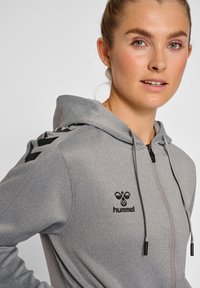 Felpa grigia con cappuccio e chiusura a zip, caratterizzata da un tessuto testurizzato, logo nero e accenti geometrici neri sulle spalle.