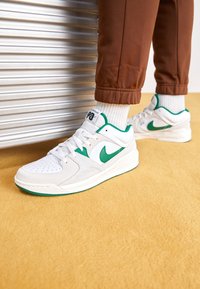 Personne portant des baskets Nike blanches et vertes avec un pantalon marron et des chaussettes blanches, debout sur un tapis jaune près d'un volet métallique.