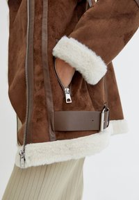 Chaqueta de piel de oveja marrón con puños de piel crema, con cremalleras plateadas, un bolsillo lateral y un cinturón marrón. Acabado de ante texturizado.