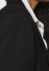 Blazer noir à texture lisse avec des épaules structurées, porté avec une chemise blanche à col visible et un détail de bouton apparent.