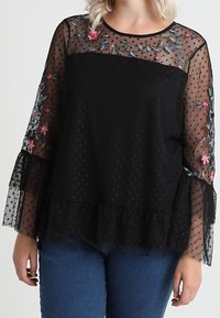 Blouse noire avec des manches transparentes à pois, ornées de broderies florales rose et bleue. Le bas présente un ourlet à volants. Associée à un jean.