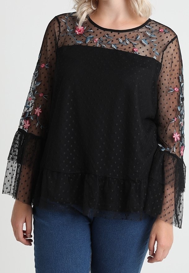 Blouse noire avec des manches transparentes à pois, ornées de broderies florales rose et bleue. Le bas présente un ourlet à volants. Associée à un jean.