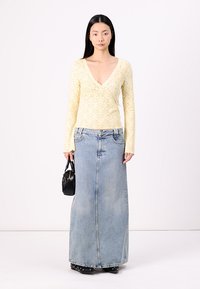 Gul spetsblus med lång ärm och V-ringning, tillsammans med en lång denimkjol och en liten svart handväska. Modell bär svarta skor.
