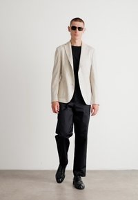 Blazer a righe di colore chiaro sopra una camicia nera, abbinato a pantaloni neri e scarpe lucide. Occhiali da sole indossati, in piedi contro uno sfondo neutro.