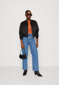 Svart bomberjacka, orange topp, högmidjade blå jeans och svarta kängor med klack. Håller en liten svart handväska med texturerad yta.