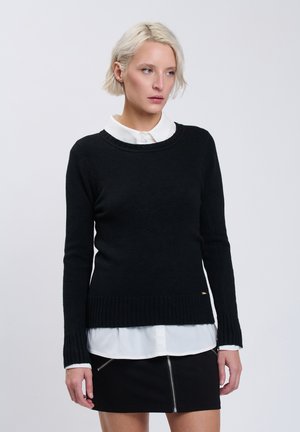 Kvind med kort blondt hår iført en sort sweater over en hvid skjorte og et sort skørt med lynlås foran, kigger til siden.