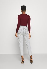 Haut bordeaux à manches longues côtelées associé à un jean taille haute gris clair. Sandales à talons noires complètent la tenue. Vue de dos, cadre minimaliste.