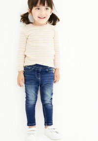 Pruhovaná dlouhá košile v světlých barvách, spárovaná s tmavě modrými skinny džíny a bílými teniskami na suchý zip. Měkká tkanina, těsný střih.
