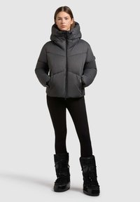 Puffedjakke i mørkegrå med høj krave, lynlåslukning og sidelommer. Sammen med sorte leggings og kraftige sorte støvler.