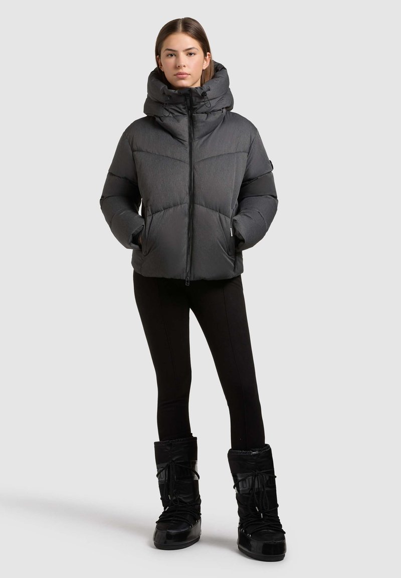 Puffedjakke i mørkegrå med høj krave, lynlåslukning og sidelommer. Sammen med sorte leggings og kraftige sorte støvler.