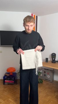 Ανοιχτόγκρι κασκόλ με φουντωτά άκρα, κρατιέται από ένα άτομο, κατασκευασμένο από μαλακό υλικό· το όνομα της μάρκας είναι ορατό κατά μήκος της κάτω άκρης.