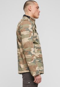 Jachetă de camuflaj în țesătură verde, maro și bej. Dispune de un guler înalt, buzunare cu nasturi și o croială lejeră cu detalii vizibile de cusături.