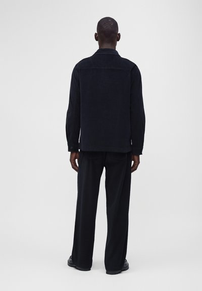 Homme portant une veste noire ample et un pantalon, debout de dos à la caméra sur un fond clair uni.
