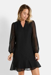Vestido negro con largas mangas abullonadas y transparentes, escote en V, textura de lunares y dobladillo acampanado. Tela ligera con una silueta fluida.