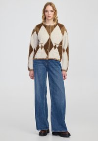 Braun- und cremefarbener Cardigan mit Rautenmuster, langen Ärmeln, vorne mit Knöpfen und zwei Taschen, kombiniert mit weit geschnittenen blauen Jeans.