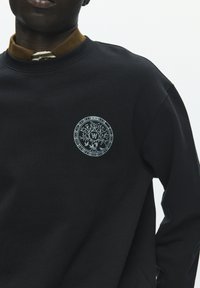 Sweat-shirt noir avec un motif rond bleu clair représentant un arbre et un logo sur la poitrine. Matière en coton, col rond, surface texturée.
