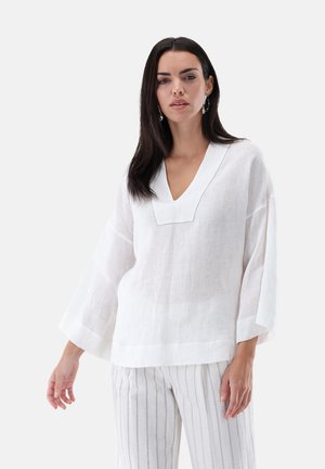 Frau trägt eine locker sitzende weiße Leinenbluse mit V-Ausschnitt und weiten Ärmeln sowie weiße Hosen mit dünnen vertikalen Streifen vor einem einfarbigen Hintergrund.