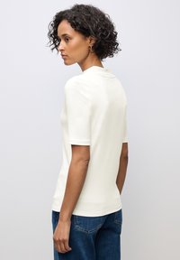 Top corto a maniche corte di colore crema con una texture liscia e un colletto sottile, abbinato a pantaloni di jeans blu. Viene mostrata la parte posteriore, evidenziando la vestibilità.