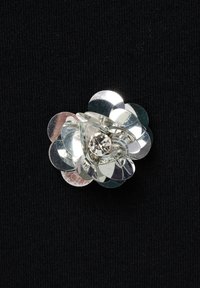 Zilveren bloem broche met gelaagde, glanzende pailletten en een centrale heldere edelsteen, tegen een zwarte gestructureerde achtergrond.