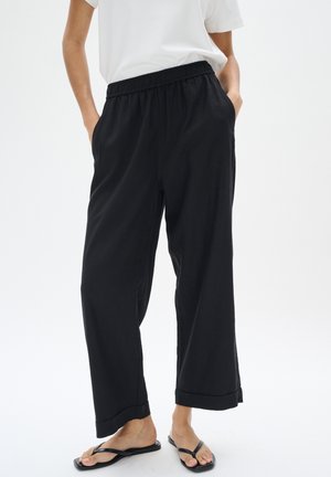 Black loose-fitting pants with an elastic waistband, side pockets, and cuffed hems, made from a lightweight fabric. Worn with black flip-flops.