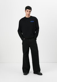 Black long-sleeve shirt with a blue logo on the chest, paired with wide-leg black pants and black shoes on a neutral background.