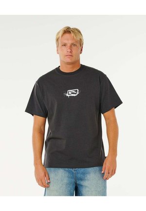 Blondyn w czarnej koszulce z małym białym nadrukiem i jasnoniebieskich dżinsach, stojący na tle jednolitego jasnego tła.