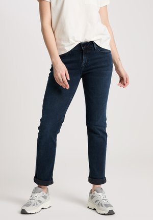 Dark blue jeans with a straight leg cut, rolled cuffs, and a classic five-pocket design, paired with a light-colored t-shirt and gray sneakers.