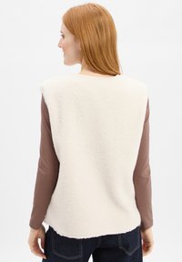 Frau mit roten Haaren, die eine ärmellose weiße Fleece-Weste über einem braunen langärmligen Oberteil und dunkelblauen Jeans trägt, von hinten gesehen.