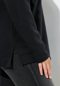 Sudadera negra con mangas largas, un corte ligeramente oversize, aberturas laterales y puños acanalados, combinada con jeans de mezclilla oscuros.