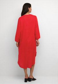 Röd linneklänning med långa ärmar, rundad halsringning och sidofickor. Buren med svarta klacksandaler, slät tygstruktur.