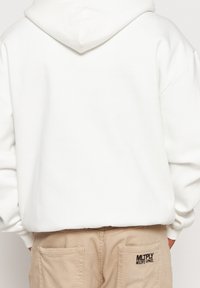 Witte hoodie met een ontspannen pasvorm, zachte textuur en een capuchon met trekkoord. Tan broek met voorzakken, voorzien van een zwart geborduurd logo.