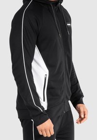 Sudadera con cremallera negra y panel lateral blanco, con capucha ajustable y un bolsillo con cremallera. Fabricada con un tejido suave y elástico.