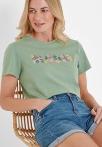 Light green cotton t-shirt featuring "BLOOM" printed in colorful floral patterns. Paired with denim shorts, sitting on a woven chair.
